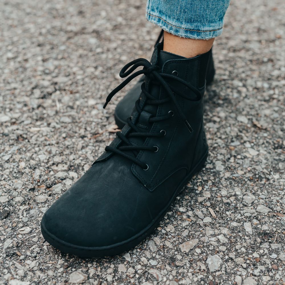 A photo of the Shapen Urbaneer combat boots made with nubuck leather, a thin wool lining, and rubber soles. The boots are black in color and have an elasticized panel on the top back of the boot. The left  boot is shown on a woman’s foot from the front left, with a view of her shin down. The woman is wearing cropped blue jeans and is standing on an asphalt road. #color_black