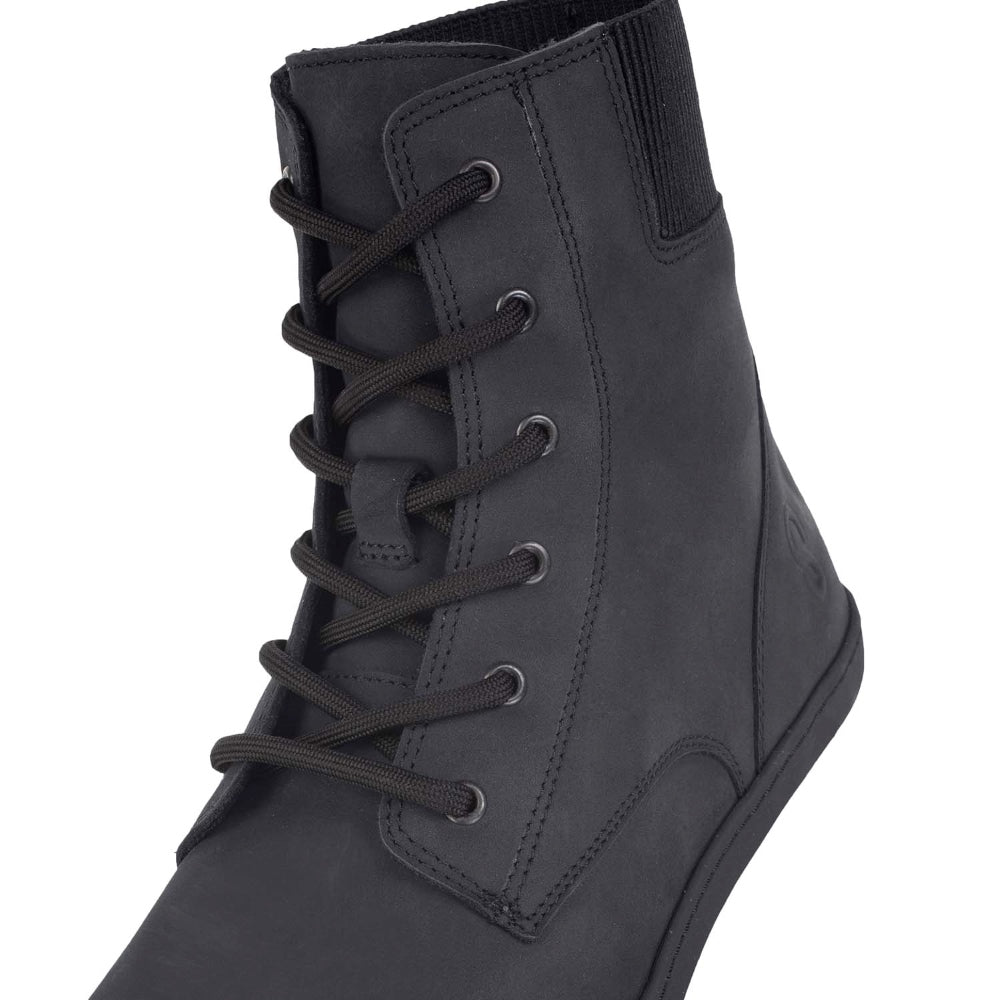 A photo of the Shapen Urbaneer combat boots made with nubuck leather, a thin wool lining, and rubber soles. The boots are black in color and have an elasticized panel on the top back of the boot. The right boot is shown from the front zoomed in on the laces, on white background. #color_black