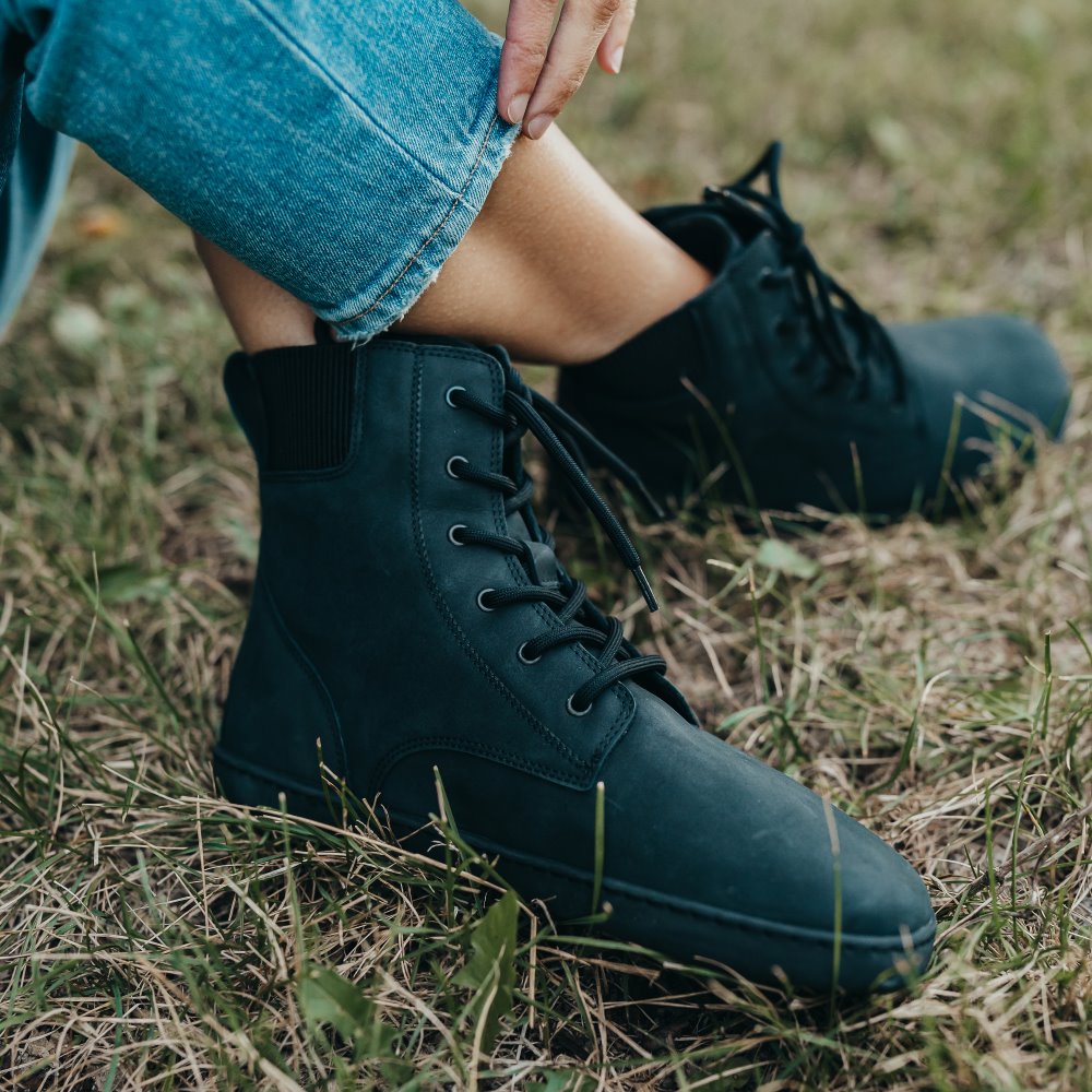 A photo of the Shapen Urbaneer combat boots made with nubuck leather, a thin wool lining, and rubber soles. The boots are black in color and have an elasticized panel on the top back of the boot. Both boot are shown on a woman’s feet from the right, with a view of her shins down. The woman is wearing cropped blue jeans and is sitting in grass. #color_black