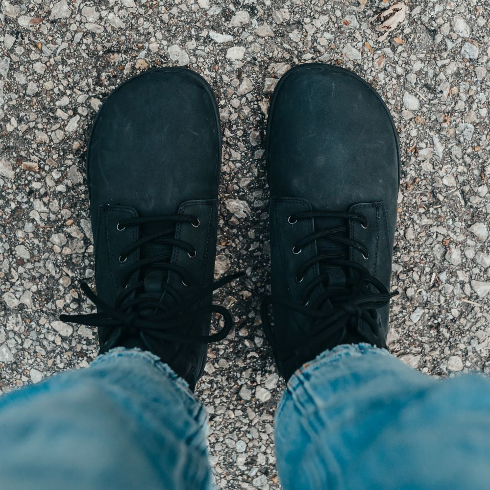 A photo of the Shapen Urbaneer combat boots made with nubuck leather, a thin wool lining, and rubber soles. The boots are black in color and have an elasticized panel on the top back of the boot. Both boots are shown from above on a woman’s feet with a view of her shins down. The woman is wearing cropped blue jeans and is standing on an asphalt road. #color_black
