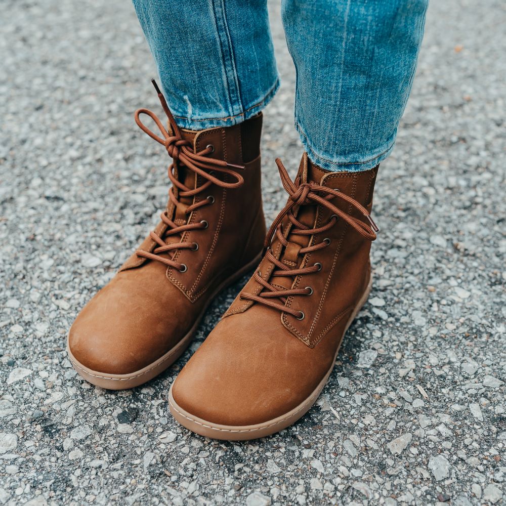 A photo of the Shapen Urbaneer combat boots made with nubuck leather, a thin wool lining, and rubber soles. The boots are caramel brown in color and have an elasticized panel on the top back of the boot. Both boots are shown from the front left  on a woman’s feet with a view of her shins down. The woman is wearing cropped blue skinny jeans with the boots, and she is standing on an asphalt road. #color_caramel