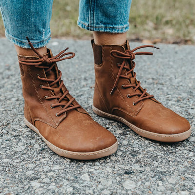 A photo of the Shapen Urbaneer combat boots made with nubuck leather, a thin wool lining, and rubber soles. The boots are caramel brown in color and have an elasticized panel on the top back of the boot. Both boots are shown from the front right on a woman’s feet with a view of her shins down. The woman is wearing cropped blue skinny jeans with the boots, and she is standing on an asphalt road. #color_caramel