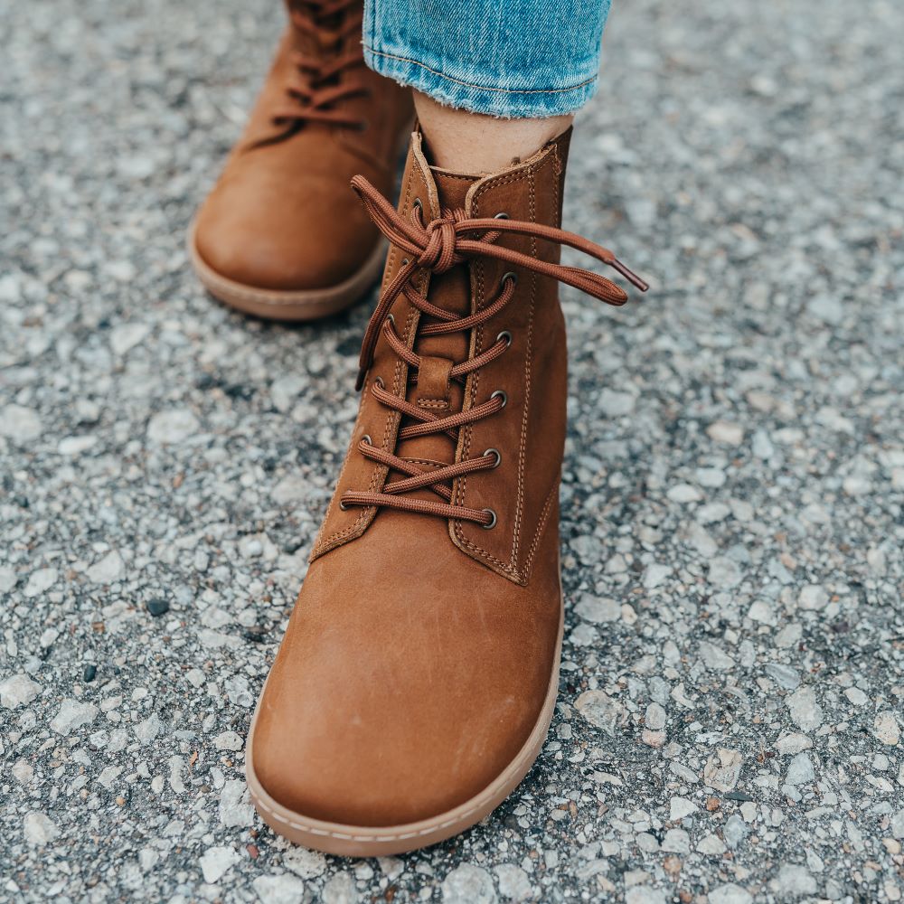A photo of the Shapen Urbaneer combat boots made with nubuck leather, a thin wool lining, and rubber soles. The boots are caramel brown in color and have an elasticized panel on the top back of the boot. Both boots are shown from the front on a woman’s feet with a view of her shins down. The woman is wearing cropped blue skinny jeans with the boots, and she is standing on an asphalt road with her left foot in front of her right. #color_caramel