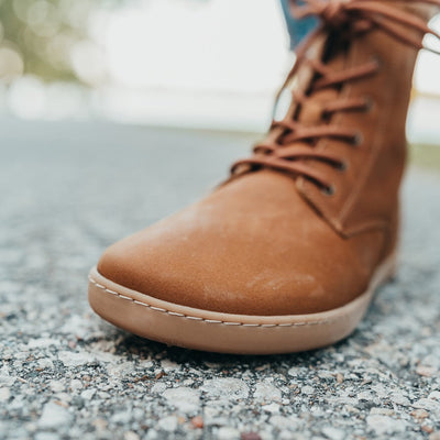 A photo of the Shapen Urbaneer combat boots made with nubuck leather, a thin wool lining, and rubber soles. The boots are caramel brown in color and have an elasticized panel on the top back of the boot. The left boot is shown from the front on a woman’s feet with a zoomed in view of the toe. The woman is standing on an asphalt road. #color_caramel