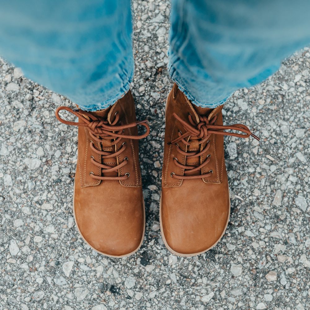 A photo of the Shapen Urbaneer combat boots made with nubuck leather, a thin wool lining, and rubber soles. The boots are caramel brown in color and have an elasticized panel on the top back of the boot. Both boots are shown from above on a woman’s feet.. The woman is wearing cropped blue jeans and is standing on an asphalt road. #color_caramel