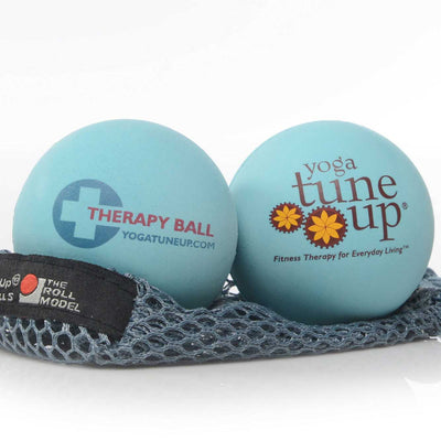 A photo of Tune Up therapy balls made from rubber. The therapy balls are a light blue color with therapy ball written on one side and yoga tune up on the other. Both balls are shown sitting on a gray mesh bag against a white background.  