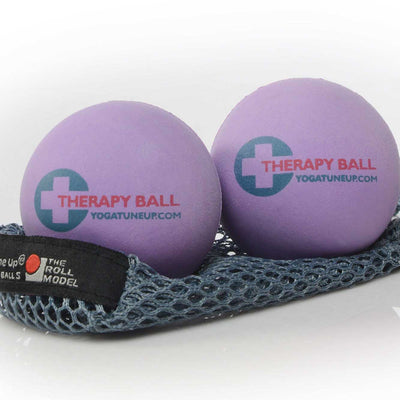 A photo of Tune Up therapy balls made from rubber. The therapy balls are a light purple color with therapy ball written on one side and yoga tune up on the other. Both balls are shown sitting on a gray mesh bag against a white background. 