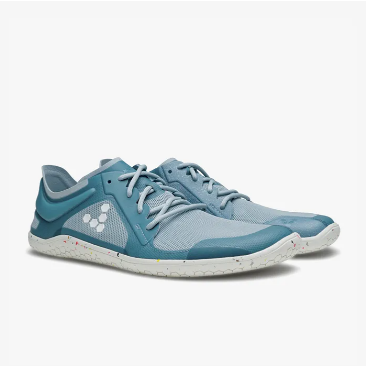A photo of Vivobarefoot Primus lite sneakers made from recycled plastic and a rubber sole. The athletic sneakers are a blue haze color with white soles spotted with color and the vivobarefoot logo on the side. Both shoes are shown beside each other from the right side angled slightly from the front against a white background. #color_blue-haze