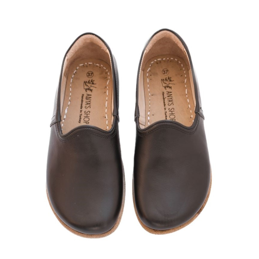 A photo of Yasemin Leather loafers Designed by Anya with a leather upper and tan rubber soles. The loafers are a black color with a smooth leather upper and have a small curve up on the top of the foot for design. Both loafers are shown from the top facing down against a white background. #color_black-smooth-leather
