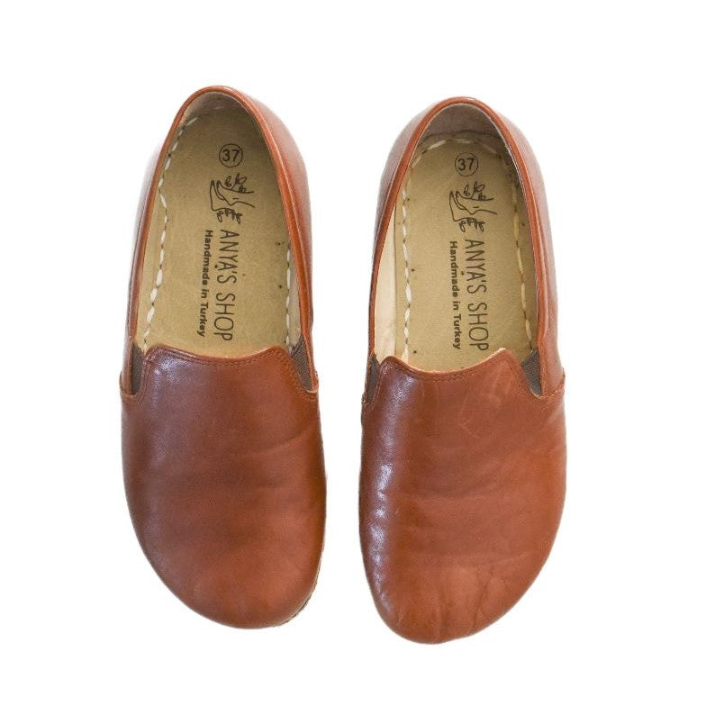 A photo of Yemeni Leather Loafers Designed by Anya with a leather upper and soles. The loafers are a brown color with tan leather soles they have small slits on each side with elastic on the top of the foot. Both loafers are shown from the top facing down against a white background. #color_brown