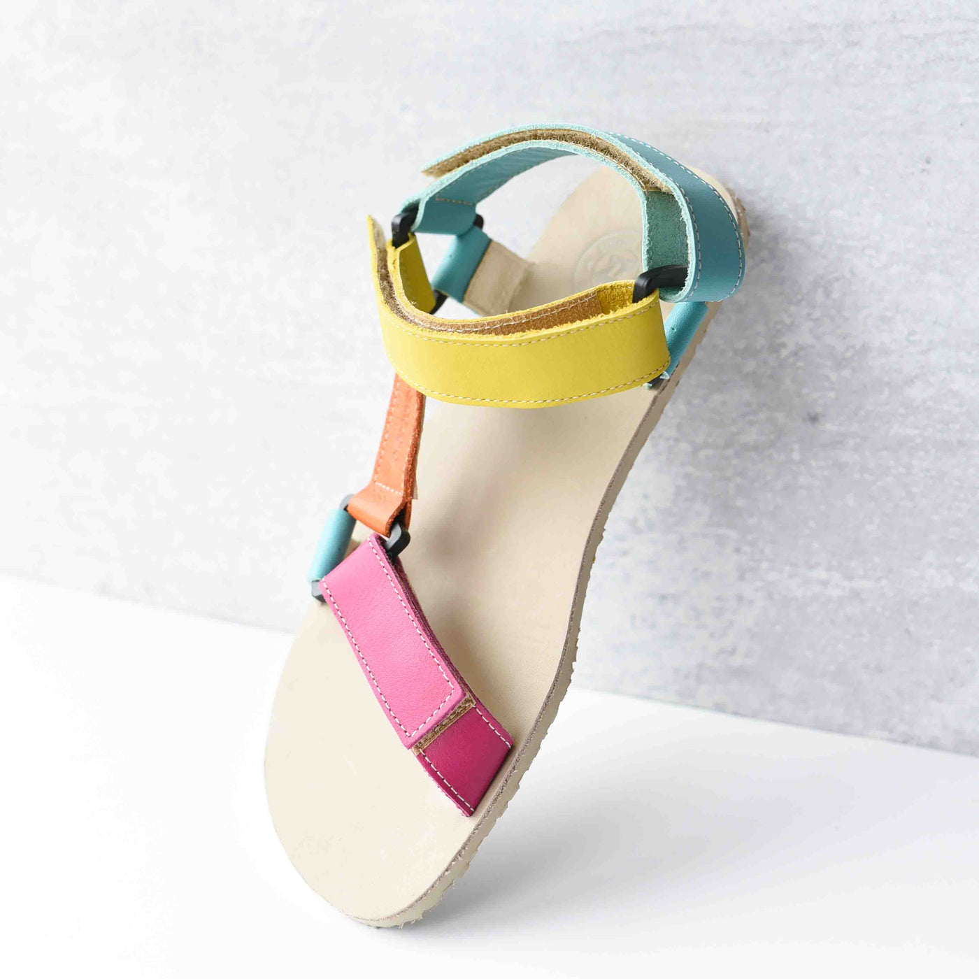 A photo of Zeezoo Olymp Rainbow sandals made from leather with velcro and a light beige leather topped rubber soles. The sandal straps are pink, light blue, orange, and yellow. The straps cross the front of the foot and a single strap runs along the outside of the top of the foot and around the ankle and heel. The left sandal is shown upright leaning up against a light grey colored wall and white floor. #color_rainbow-olymp
