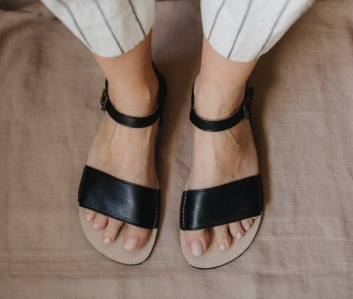 A photo of Zeezoo Siren Black Sandals made of leather and a light beige leather topped black rubber sole. The sandals have a front foot crossing strap, a leather ankle strap and heel cup. A woman is shown from mid leg down wearing white black striped pants and the sandals standing straight to the front on a tan sheet on the floor. #color_black