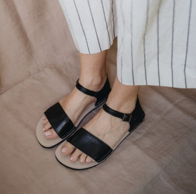 A photo of Zeezoo Siren Black Sandals made of leather and a light beige leather topped black rubber sole. The sandals have a front foot crossing strap, a leather ankle strap and heel cup. A woman is shown from knee down wearing white black striped pants and the sandals standing to the side on a tan sheet on the floor. #color_black